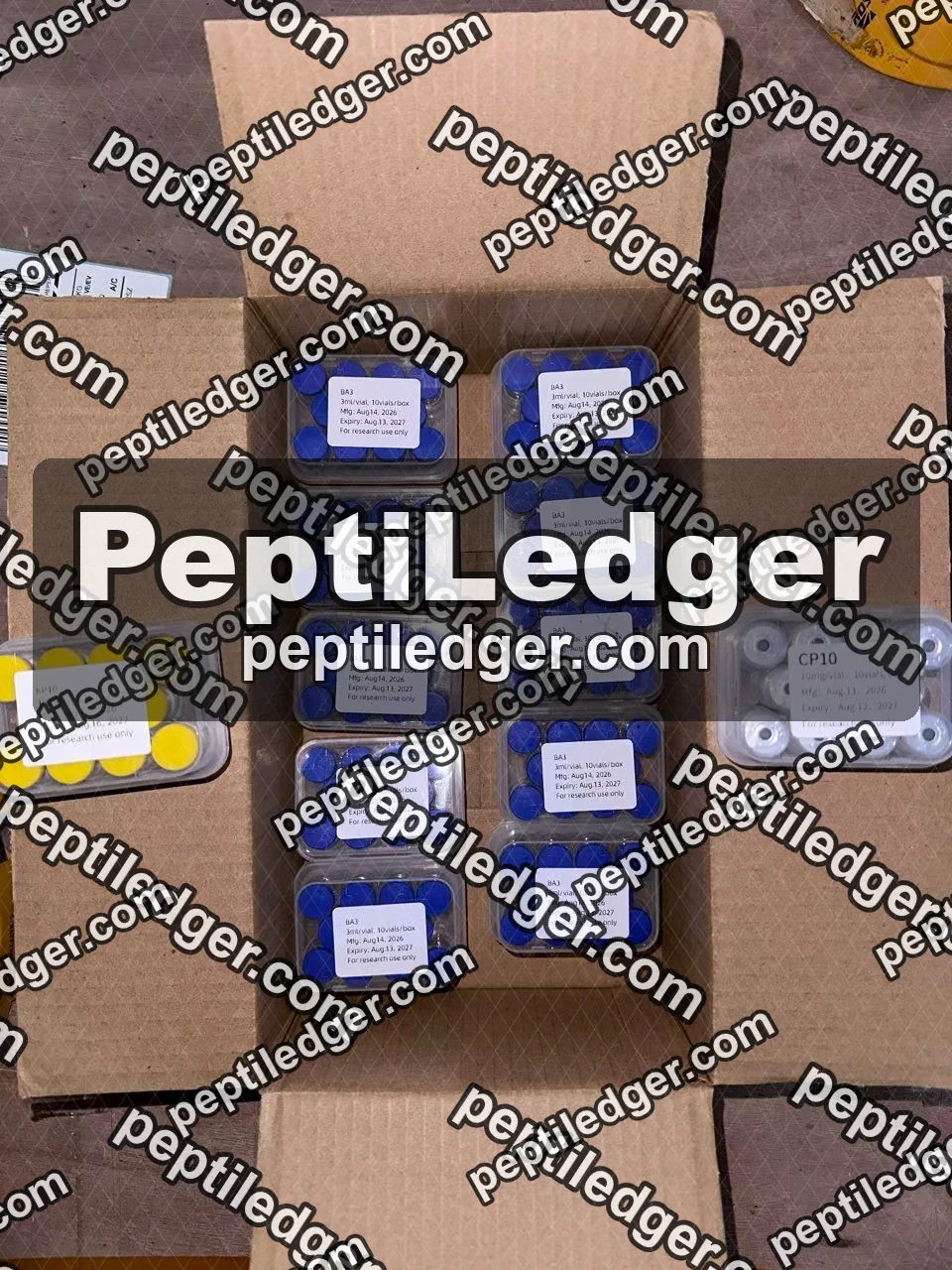 An open cardboard carton with ten boxes of blue-capped vials standing in two rows, lid labels facing up, with two further boxes of different codes beside the carton.